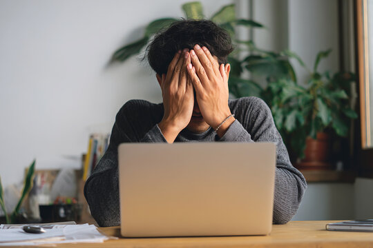 Exhausted woman covering her face with hands sitting in front of a laptop on a work desk at home against a background of indoor plants