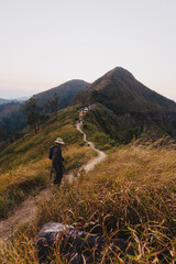 Obraz premium A hiker walks along a narrow dirt trail on a grassy mountain ridge heading towards a scenic peak at twilight.