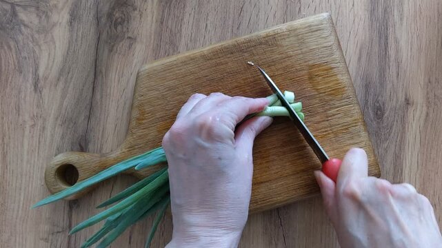 Female hands chopping fresh green onions with a sharp knife on a wooden board. Sliced scallion pieces stand on a cutting desk. Healthy food cooking concept