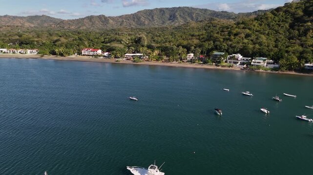 Aerial drone video of Playa Potrero on Costa Rica's Pacific coast in Guanacaste Province. Tropical bay with anchored boats, beachfront homes, lush forest-covered hills, and a scenic shoreline under cl