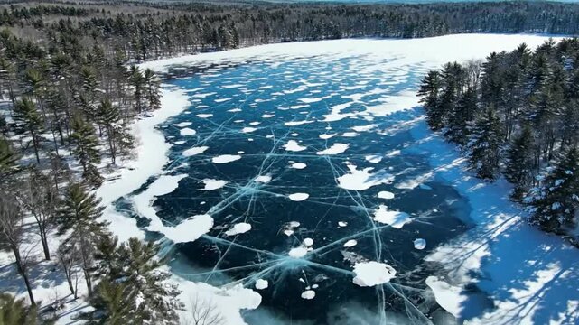 Aerial view of a frozen lake with ice cracks and snowy shoreline surrounded by evergreen trees on a sunny winter day