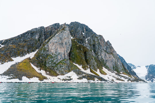 View of a massive rocky mountain with patches of snow and green moss rising from the arctic sea with small red boats near the shore in Svalbard and Jan Mayen.