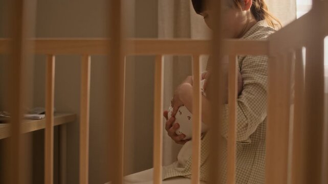 Tilt up shot of young Caucasian mother on maternity leave sitting at table with laptop and financial documents and rocking infant girl in arms in cozy nursery room with wooden crib in foreground