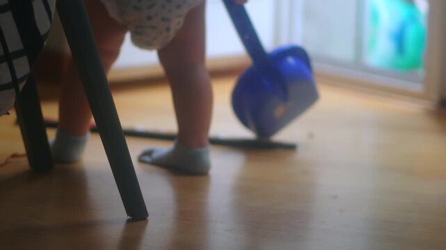 Toddler walking indoors while carrying blue toy shovel before crouching down and placing it on floor capturing everyday curiosity, movement learning and authentic childhood behavior at home