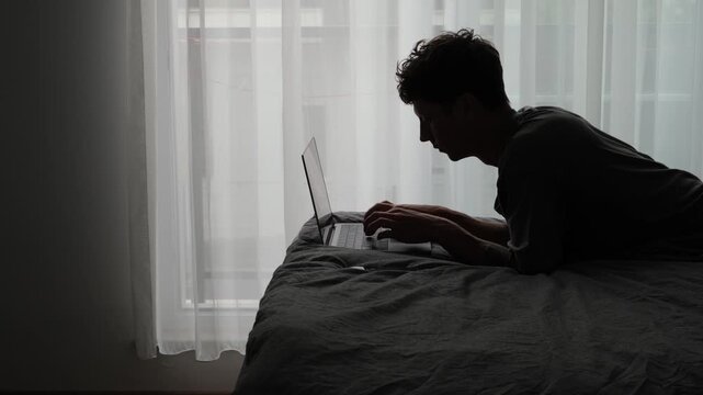 Silhouette caucasian man typing on laptop by window, moody bedroom scene with curtain backlight, freelancer working late, hunched posture, introspective focus