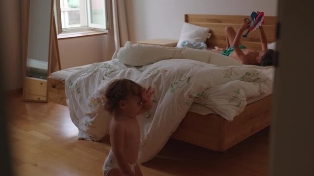 Toddler walking through bedroom toward older sibling playing on bed with toy blasters capturing everyday family interaction, curiosity and authentic childhood home life