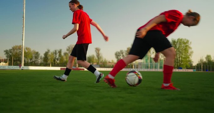 Young soccer player in red jersey practicing footwork on a green outdoor field at sunset, with ball, goal area, trees, and stadium lights