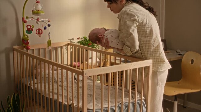 Medium long shot of caring young Caucasian mother entering cozy nursery room and putting her infant daughter in crib for afternoon sleep