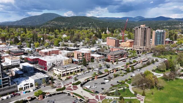 Coeur d'Alene downtown with modern high-rises, charming brick storefronts, green parks, construction crane and dense pine forest stretching toward blue mountain ranges in spring. Aerial wide shot.