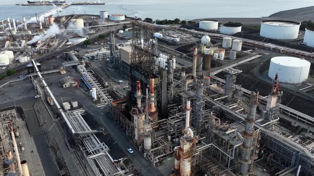 Large oil refinery in Rodeo California with complex piping systems and white storage tanks near San Pablo Bay with smoke rising from industrial stacks during overcast daytime in USA. Aerial flyover.