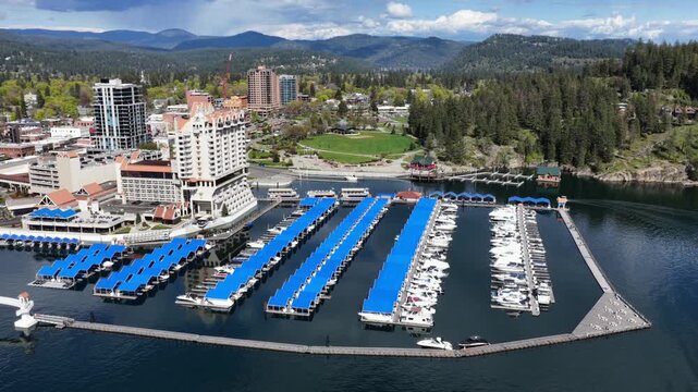 Coeur d Alene, Idaho resort and marina with blue boat slips on lake with downtown buildings and lush green pine trees under bright blue sky in Pacific Northwest United States. Aerial orbit wide shot.