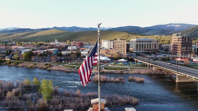 Missoula downtown with American flag, Clark Fork River, bridge, riverfront park, brick buildings and rolling hills with snow-capped mountain range under clear sunny sky.Montana, USA. Aerial orbit view