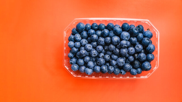 Fresh blueberries filling a clear plastic punnet, offering a vibrant contrast against the solid orange background, highlighting concepts of healthy eating and fresh produce