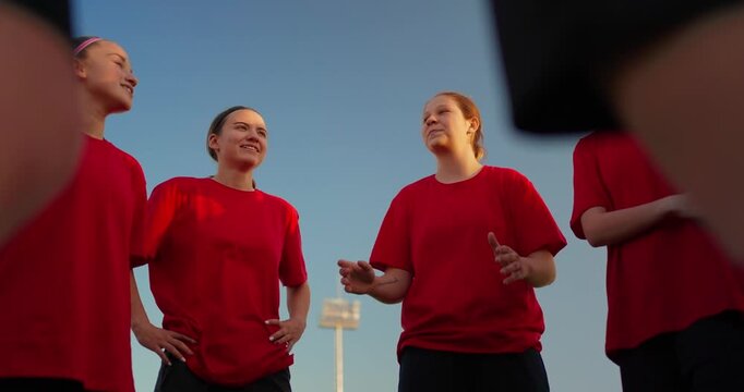 Low angle view of young girls in red jerseys smiling together in a team huddle on an outdoor sports field under a clear evening sky