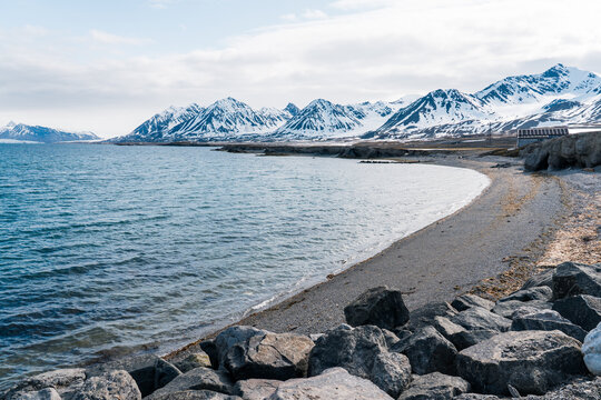 View of the rocky coastline and calm blue waters with snow-capped mountains under a bright sky in Ny-Alesund, Svalbard, Svalbard and Jan Mayen.