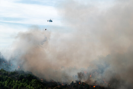 Opera&ccedil;&atilde;o de combate a um inc&ecirc;ndio florestal com um helic&oacute;ptero bombeiro a executar a descarga de &aacute;gua com um balde bambi em meio a uma densa coluna de fumo sobre uma encosta com focos ativos de fogo