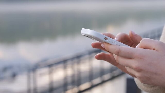 Close-up of hands holding a white smartphone outdoors. Person stands near railing overlooking water. Background shows soft-focused river and railing. Light reflects softly on phone and skin