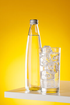 Glass of sparkling water with ice cubes on yellow background minimal