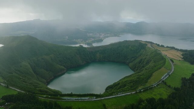 Breathtaking aerial view of the Sete Cidades volcanic caldera, showcasing the vibrant green crater lake in the foreground and the expansive twin lake system in the background, set against misty