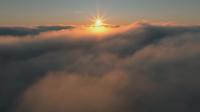 Breathtaking aerial view above a dense layer of clouds at sunset, with the sun casting a golden glow and starburst effect over the softly illuminated cloudscape.