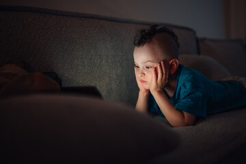 Obraz premium Child watching glowing digital tablet screen in dark home, concentrating on online content, technology addiction concept