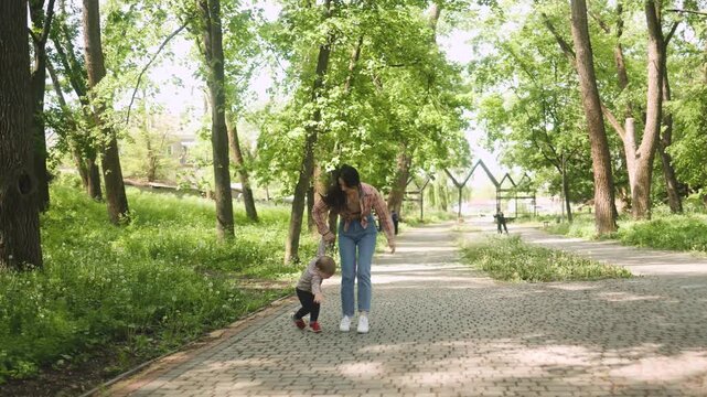 A young beautiful mother holds her little son's hand as he takes playful steps along a paved path in a sunny green park. Family, motherhood, childhood, spring, walk, nature, happiness, togetherness