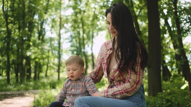 A young beautiful mother gently embraces her little son and pats his head while sitting together in a lush green public park. Family, motherhood, love, childhood, togetherness, care, happiness