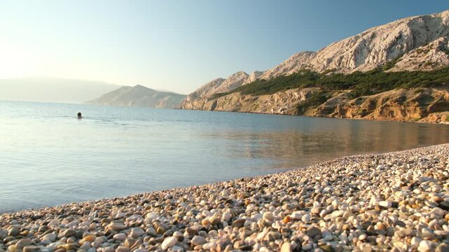 man swimming Calm pebble beach with sea and rocky mountains