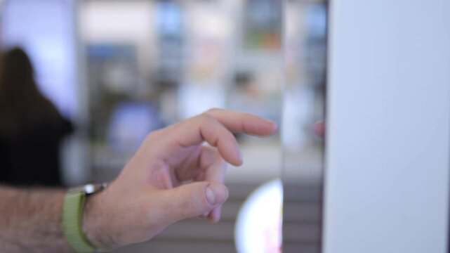 Hand taps screen, focusing on interaction. Arm has watch and green wristband. Background blurred with people moving. Shows modern touch interface. Good for tech, apps, or user engagement clips