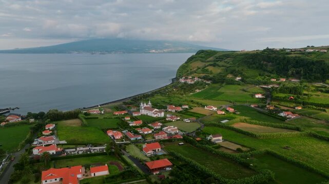 Aerial view of a picturesque coastal village on Faial Island in the Azores, featuring white houses with orange-red roofs set against vibrant green pastures, with the dramatic coastline and the distant
