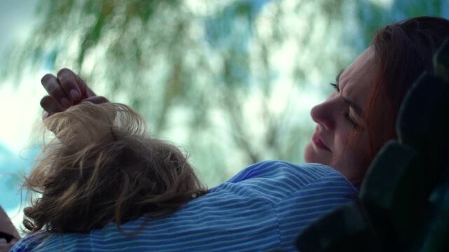 Mother softly stroking toddler hair while child relaxes against her chest outdoors capturing warmth, reassurance and authentic parent child connection on peaceful summer day