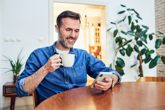 Man smiling at smartphone with coffee at home dining table