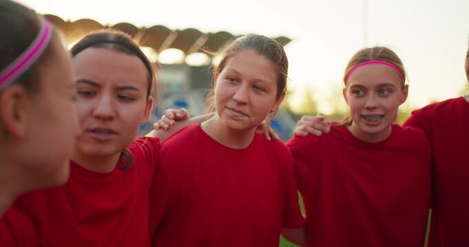 Young female soccer players in red jerseys sharing a supportive team huddle on the field at sunset, smiling with stadium seats behind