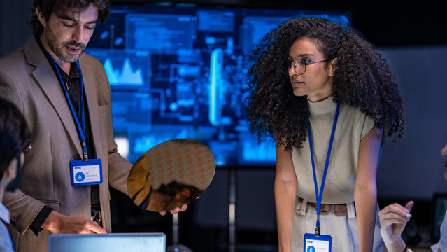 A multiethnic technology developer team holding and inspecting a raw silicon wafer disk to check semiconductor circuit microchip fabrication logic parameters in a workspace.