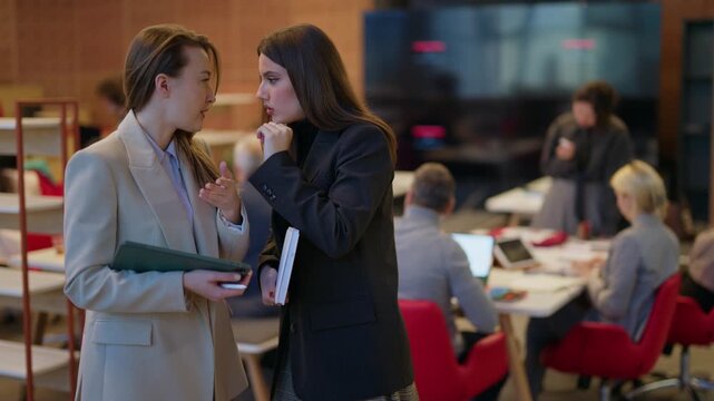 Two colleagues share a private moment, one whispering into the other's ear. Employees engage in a confidential conversation within a bustling workplace.