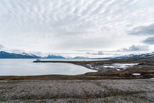 View of the historic Bamsebu hut on a coastal spit with a ship in the fjord and snow-capped mountains under a cloudy sky Svalbard, Svalbard and Jan Mayen.