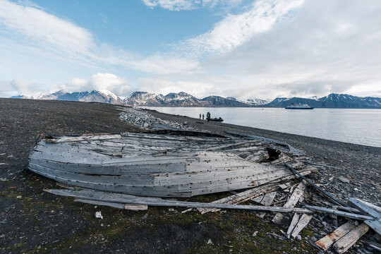 View of the remains of an old wooden boat at Bamsebu on a rocky beach with whale bones, people and snow-capped mountains in Svalbard, Svalbard and Jan Mayen.