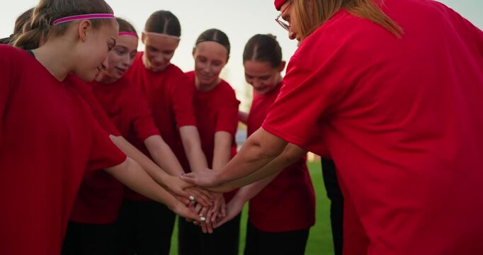 Young female soccer team in red shirts huddling with hands together and applauding on a grassy field during an outdoor practice at sunset