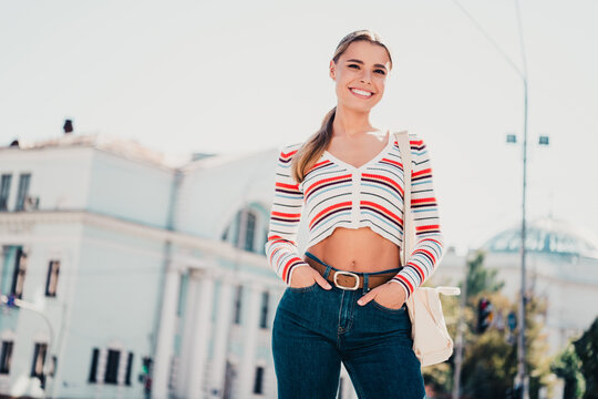 Smiling young woman in striped crop sweater and jeans posing confidently outdoors in a bright urban setting, casual fashion, summer street style and positive lifestyle portrait