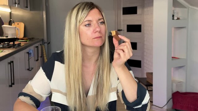 Blonde White Woman Nibbling Chocolate Bar And Sipping Water At Kitchen Table Tasting Sweet Bite Glass In Hand