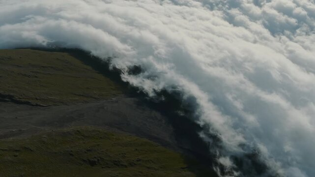 Aerial view of a dense blanket of clouds spilling over the rugged, dark volcanic slopes of Mount Pico, Azores, creating a dramatic contrast between the earth and the sky.
