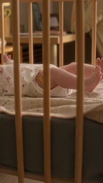 Vertical cropped shot of unrecognizable newborn baby lying in wooden crib in modern nursery room with mother sitting at table nearby