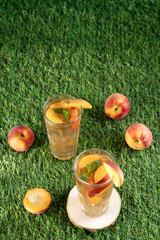Naklejka premium High angle view of two glasses of cold peach tea with fresh mint, scattered ripe peaches around on a green grass lawn