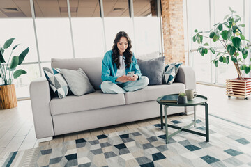 Obraz premium Young woman relaxing on a sofa in a bright living room using a smartphone, cozy home interior with plants, coffee table and decorative pillows