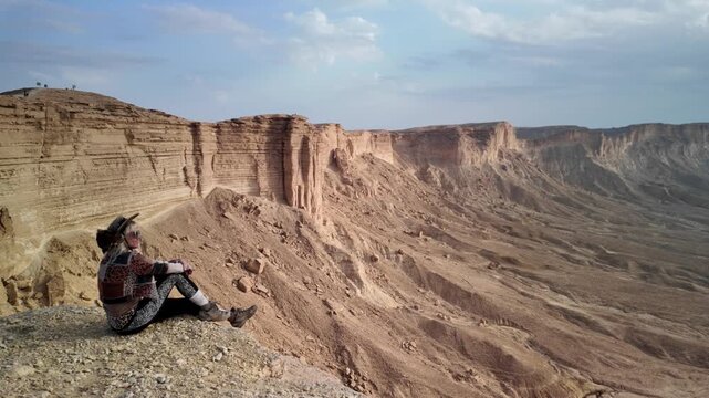 woman tourist sitting on the edge of the world escarpment at sunset, contemplating the cliffs of Tuwaiq mountain range in Riyadh, Saudi Arabia, a breathtaking natural wonder