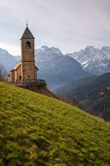 Fototapeta premium Historic stone church on a green hill with snowy Dolomite mountain peaks in Casamazzagno, Italy