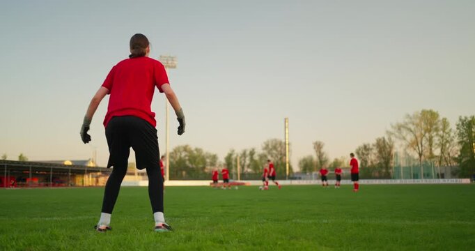 Soccer player in red training kit warms up then dives onto a green practice field as teammates run drills during an outdoor evening session