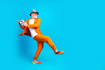 Naklejka premium Senior man in bright orange suit and hat holding a disco ball while dancing against a blue background — playful fashion, retro party and energetic movement