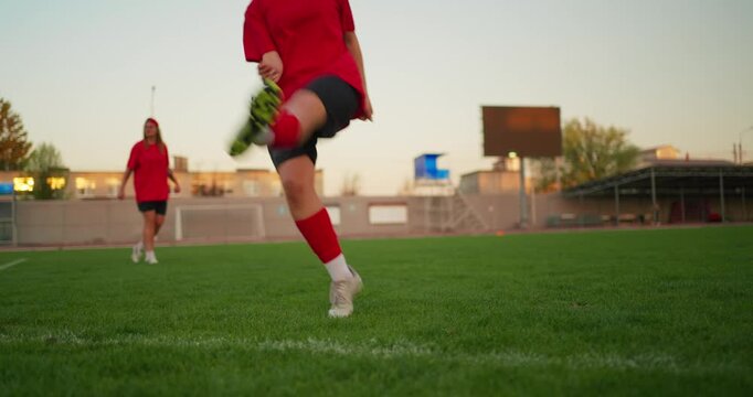 Young soccer players in red uniforms practice passing on a green field at sunset, with a ball in focus near the sideline and stadium backdrop