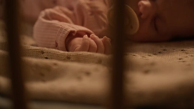 Cropped closeup shot of tiny hand of adorable newborn with pacifier in mouth lying in crib on soft blanket and sleeping peacefully at night